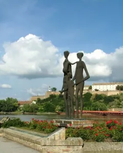 Monument to the Victims of Fascism with bronze figures on a brick platform beside flowering bushes and a river.