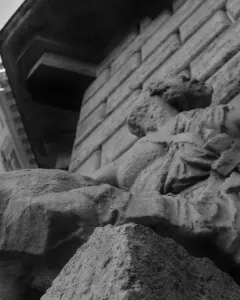 Weathered Pasquino stone statue with eroded draped figures beside a stone building and a metal rod protruding from the shoulder.