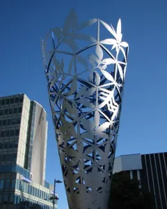 The Chalice metal sculpture rising against a blue sky in Christchurch, with modern buildings behind it.
