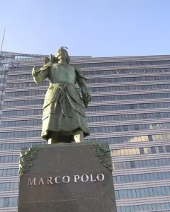 Marco Polo statue in historical attire with a bird on his arm, standing on a stone pedestal.