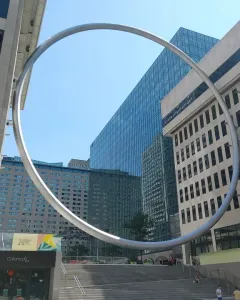 The Ring sculpture curves above a public staircase between tall city buildings.