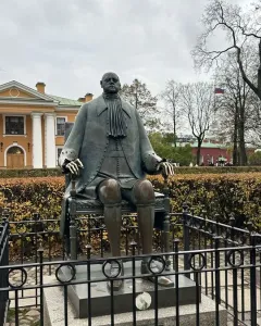 Peter I Monument shown as a bronze statue of a seated man on a stone pedestal with a low fence around it.