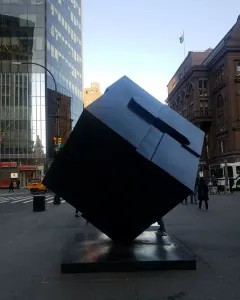 The Alamo sculpture shown as a large dark cube tilted on one corner in an urban plaza.