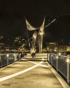 Life Electric sculpture lit at night on a waterfront promenade with Como city lights and a dark mountainside in the background.
