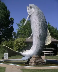The Big Trout sculpture standing in a circular pool with trees and a gazebo nearby.