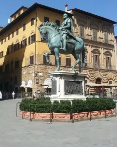 Cosimo I equestrian monument on a tall stone pedestal in a city square, with historic buildings and a few people nearby.