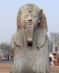 The Sphinx of Memphis, a weathered stone sphinx with a pharaonic headdress and false beard, stands on a rough base amid palm trees.