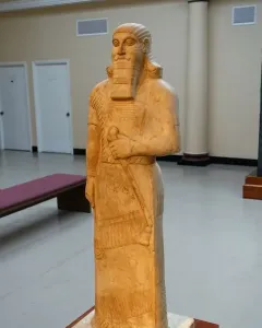 Statue of Ashurnasirpal II standing on a pedestal in a museum gallery, with a bearded figure holding an object across his chest and wearing a long patterned tunic and fringed skirt.
