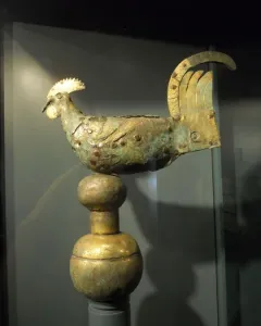 Gallo di Ramperto, a weathered metallic bird sculpture with traces of gold on its crest and tail, mounted on a tiered spherical pedestal inside a display case.