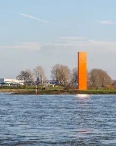 Rheinorange sculpture standing beside a wide river with sunlight reflecting on the water and distant buildings in the background.