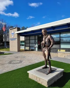 The Ideal Scout bronze statue on a stone pedestal outside a modern building with a blue sign and American flag.