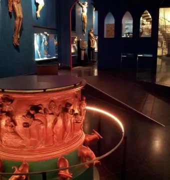 Baptismal font with reddish-brown sculpted reliefs and small animal figures around the base, displayed in a dim gallery.