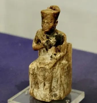 Khufu statuette of a seated figure wearing a tall headdress and shown on a clear base.