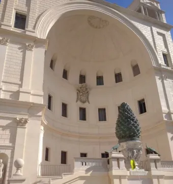 La Pigna pine cone sculpture on an ornate pedestal with peacock statues in front of a curved building facade.