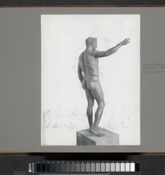 Rear-left view of the Antikythera Ephebe bronze statue, showing a nude male figure on a plinth with one arm extended.
