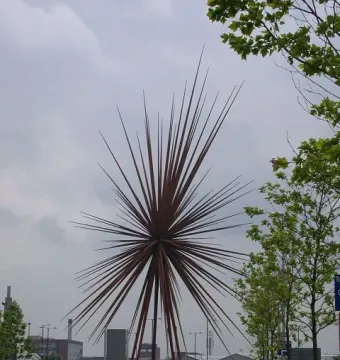 B of the Bang, a rusty-red starburst sculpture with long spikes radiating outward in a paved plaza.