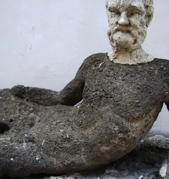 Weathered reclining figure on the Babuino Fountain, with a dark textured body and lighter bearded head.
