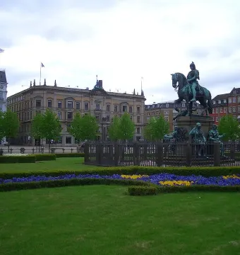 Christian V equestrian statue on a stone pedestal behind a fence in a landscaped park with flower beds and historic buildings beyond.