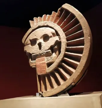 Disk of Mictlāntēcutli stone sculpture with a skull-like face and semicircular ribbed frame in a museum display.