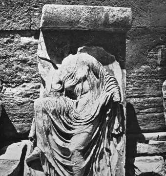 Damaged relief sculpture of Nike Untying Her Sandal, shown headless and draped in flowing robes beside a stone wall.