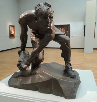 Bronze sculpture of a crouching man holding a rough stone, displayed on a pedestal in a gallery.