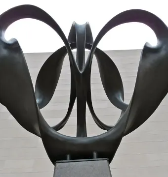 Continuum, a dark metal sculpture with upward-curving symmetrical loops on a pedestal against a stone wall, seen from below.