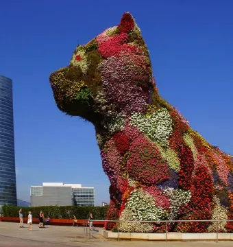 Jeff Koons’ Puppy topiary sculpture covered in flowers, with a skyscraper and city buildings behind it.