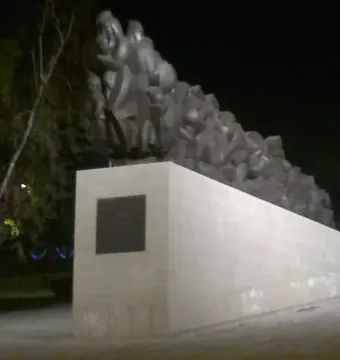 The Train of Pain monument at night, showing a long dark sculpture of clustered human figures on a light-colored base.