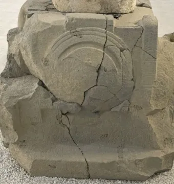 Weathered stone block with carved semicircular motifs and faint inscription lines, showing heavy cracking and damage.