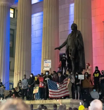 A George Washington statue stands between stone pillars as a crowd gathers below with signs and an American flag.