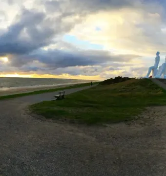 The Man by the Sea sculpture overlooking a beach and the sea at sunset, with four white seated figures on a grassy mound.