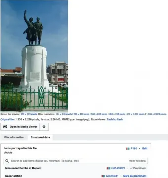 Monument Demba et Dupont with two dark figures on a tall white cylindrical base behind a green fence.