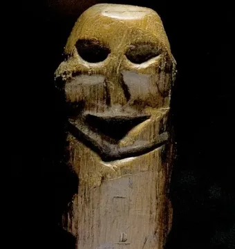 Mannetje van Willemstad wooden figure with a weathered surface and hollowed facial features on a clear base.