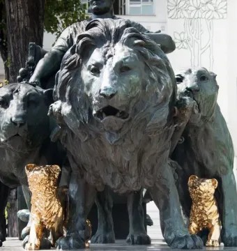 Marc Anton Group bronze sculpture with large lions and two small golden cub figures at the base, outdoors near trees and a white building.