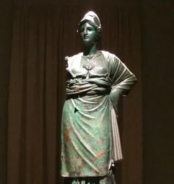 Minerva d\'Arezzo, a green-patinated armored figure standing on a wooden pedestal against a dark backdrop.