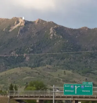 Our Lady of the Rockies statue on a distant mountain peak above the trees, with a highway sign and bridge in the foreground.