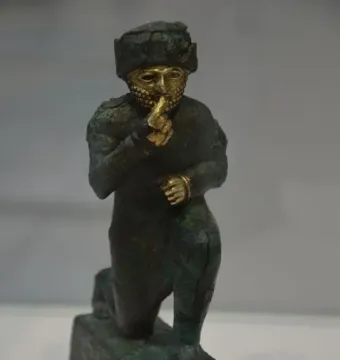 Worshipper of Larsa kneeling with one hand raised to its mouth, showing a dark bronze body and a golden face and beard.