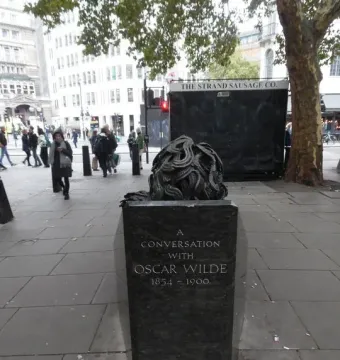 A Conversation with Oscar Wilde monument on a city sidewalk, with a dark sculptural head on top and pedestrians in the background.