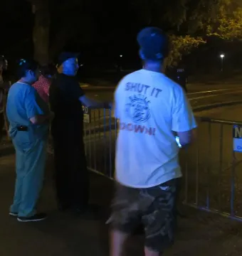 People stand near crowd control barriers at night beside a N.O.P.D. police line sign, with one person wearing a \