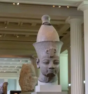 Colossal head of Amenhotep III displayed on a white pedestal in a museum gallery, with a smaller stone fragment beside it.