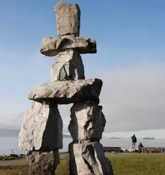 Inukshuk stone monument beside a grassy shoreline with water and distant ships under a clear blue sky.