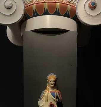 Korè 675, a brightly colored female sculpture with braided orange hair, stands beneath a painted architectural capital.