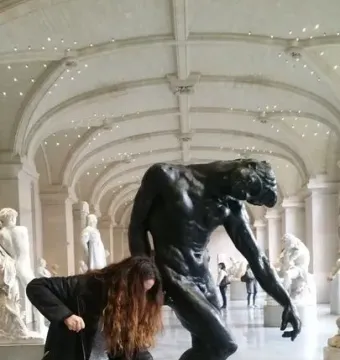 The Shade sculpture by Rodin in a museum hall, with a person posing beside it in a similar bent-forward stance.