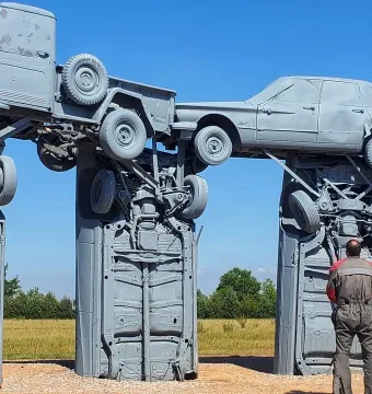 Carhenge made from grey-painted cars and trucks standing in a grassy field under a clear blue sky, with a person looking up at the structure.