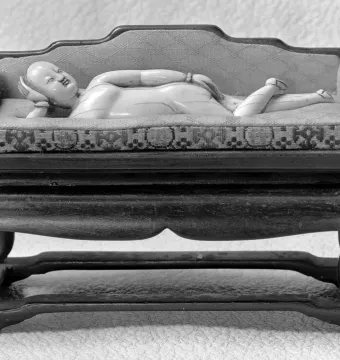 Doctor\'s Lady, a light-colored carved female figure reclining on a patterned daybed with an upholstered back.