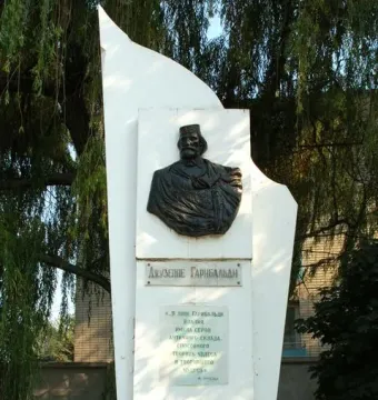 Garibaldi Monument with a dark bust relief and plaques on a white angular pedestal, surrounded by greenery.
