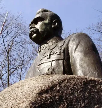 Bronze bust of Józef Piłsudski in uniform on a rough stone pedestal, with bare branches and a striped pole against a blue sky.