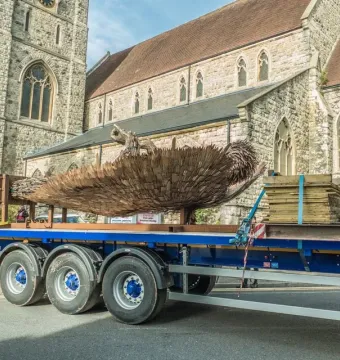 The Knife Angel sculpture леж on a blue flatbed trailer beside a road, with a stone church and clock tower in the background.