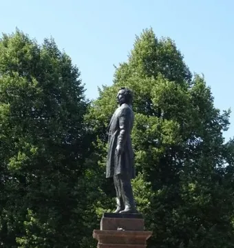 Pushkin statue on a tall pedestal in a park with trees and blue sky behind it.