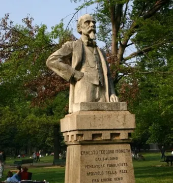 Monument to Ernesto Teodoro Moneta on a tall stone pedestal in a park, with trees and people on the lawn behind it.
